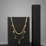 Double-layer gold necklace featuring a flat herringbone chain and a delicate chain with Tree of Life charms and gold beads on a black pedestal.