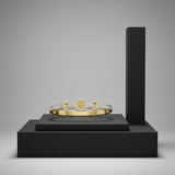 A gold bangle bracelet featuring a central circular emblem and diamond-encrusted bands, displayed on a black geometric pedestal.