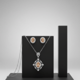 A silver or white gold necklace and earring set featuring oval-cut peach morganite gemstones surrounded by diamond halos, displayed on a black geometric stand.