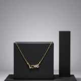 Gold chain necklace featuring a diamond-encrusted "LOVE" pendant with a heart-shaped "O" on a black display stand.