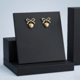 Pair of gold heart-shaped drop earrings with bow detail on a black premium display stand.