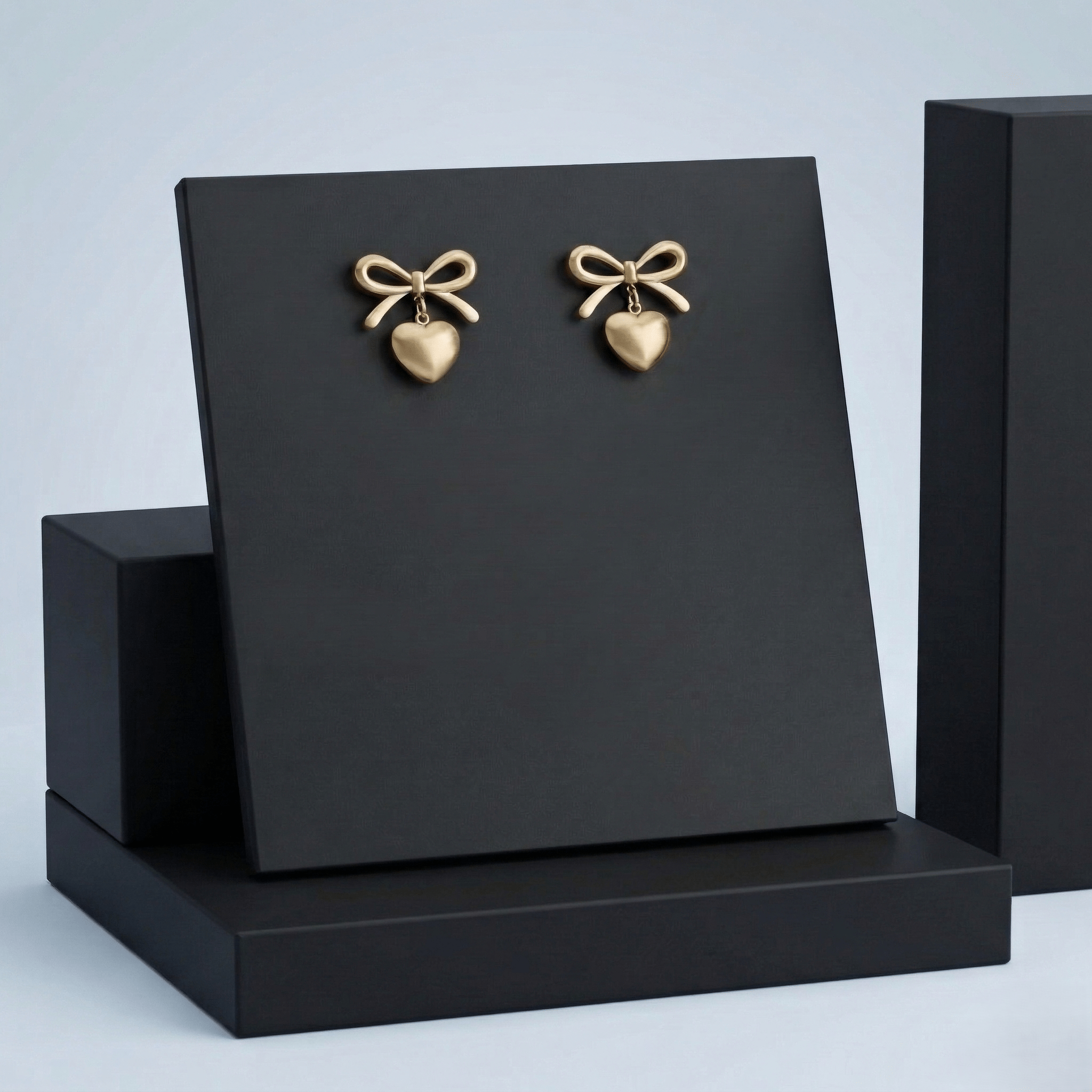 Aurelia-Gold-Heart-Bow-Earrings-Luxury Pair of gold heart-shaped drop earrings with bow detail on a black premium display stand.